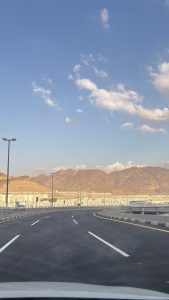 An empty curving multi-lane highway leads toward a vast field of white tents at Mina, below sunlit rocky mountains under a blue sky with scattered clouds.
