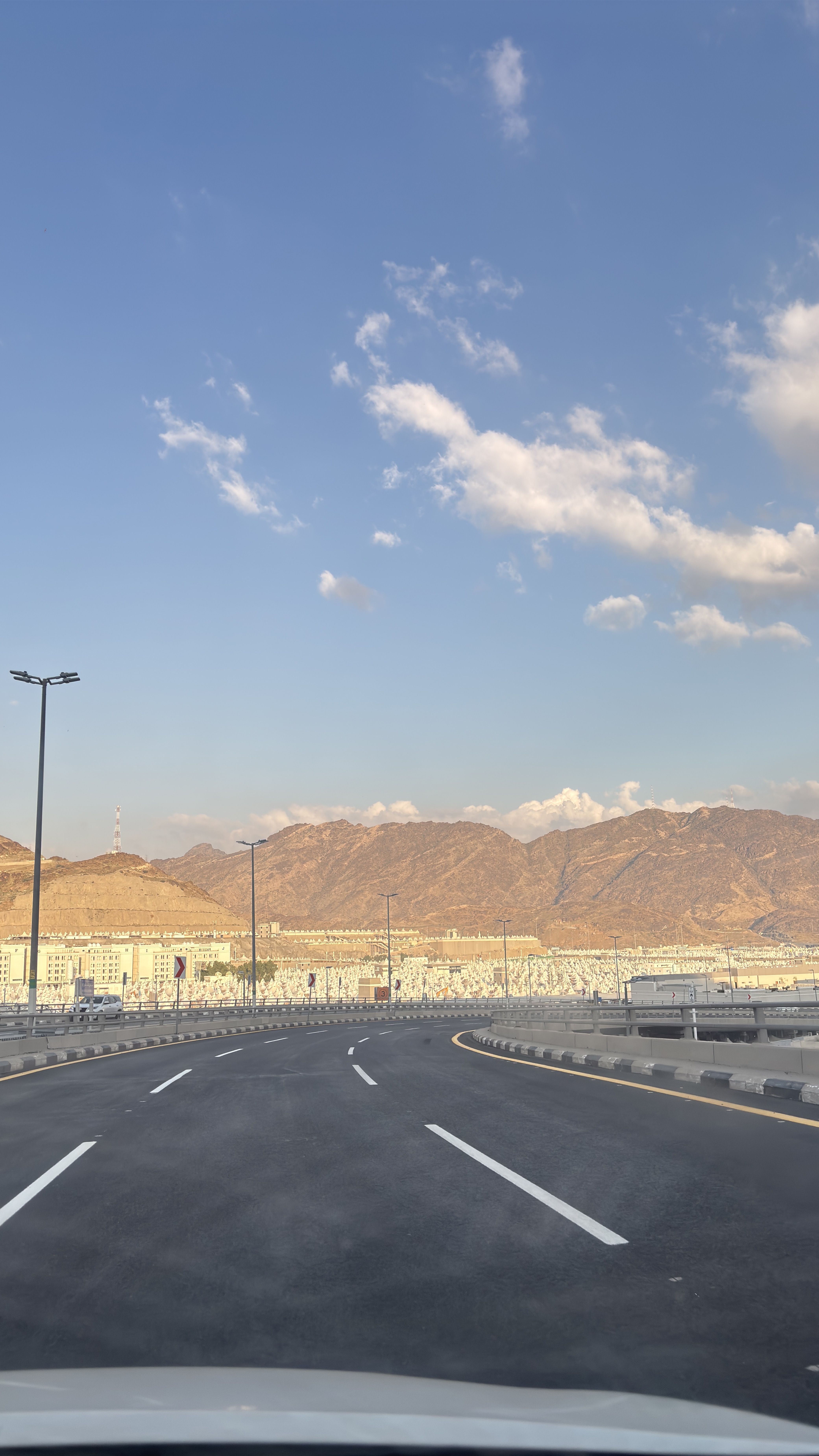 An empty curving multi-lane highway leads toward a vast field of white tents at Mina, below sunlit rocky mountains under a blue sky with scattered clouds.