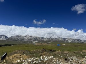 Green plateau with small tents, rugged mountains, and a layer of white clouds under a blue sky.