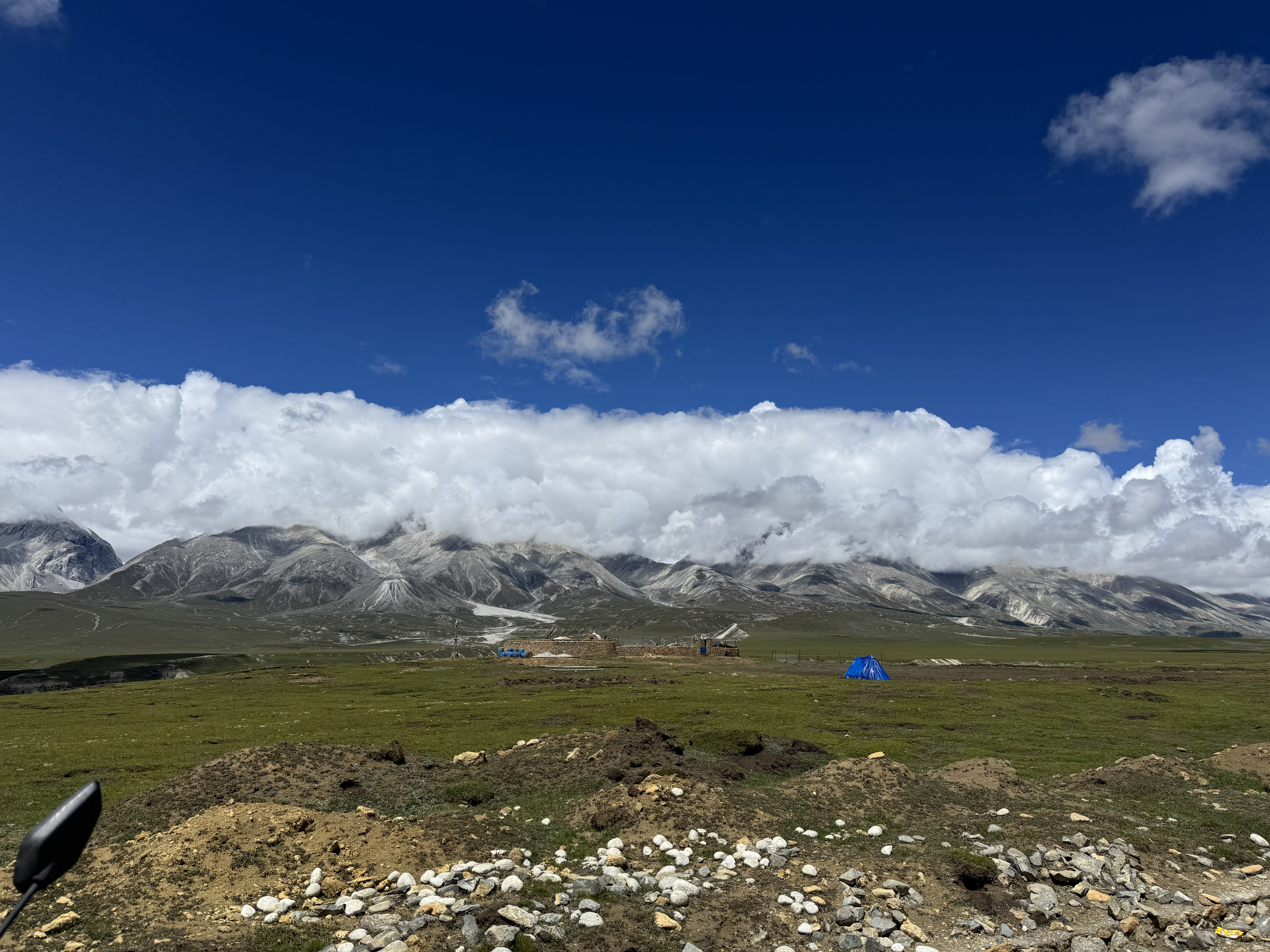 Green plateau with small tents, rugged mountains, and a layer of white clouds under a blue sky.