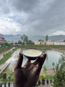 A close-up of a hand holding a traditional clay pot containing Juju Dhau (king curd), set against the blurred background of Nag Pokhari pond and surrounding hills.
