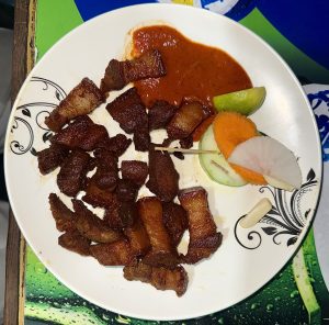 Crispy fried meat served with spicy sauce, lime slices, carrots, and cucumber on a decorative white plate.
