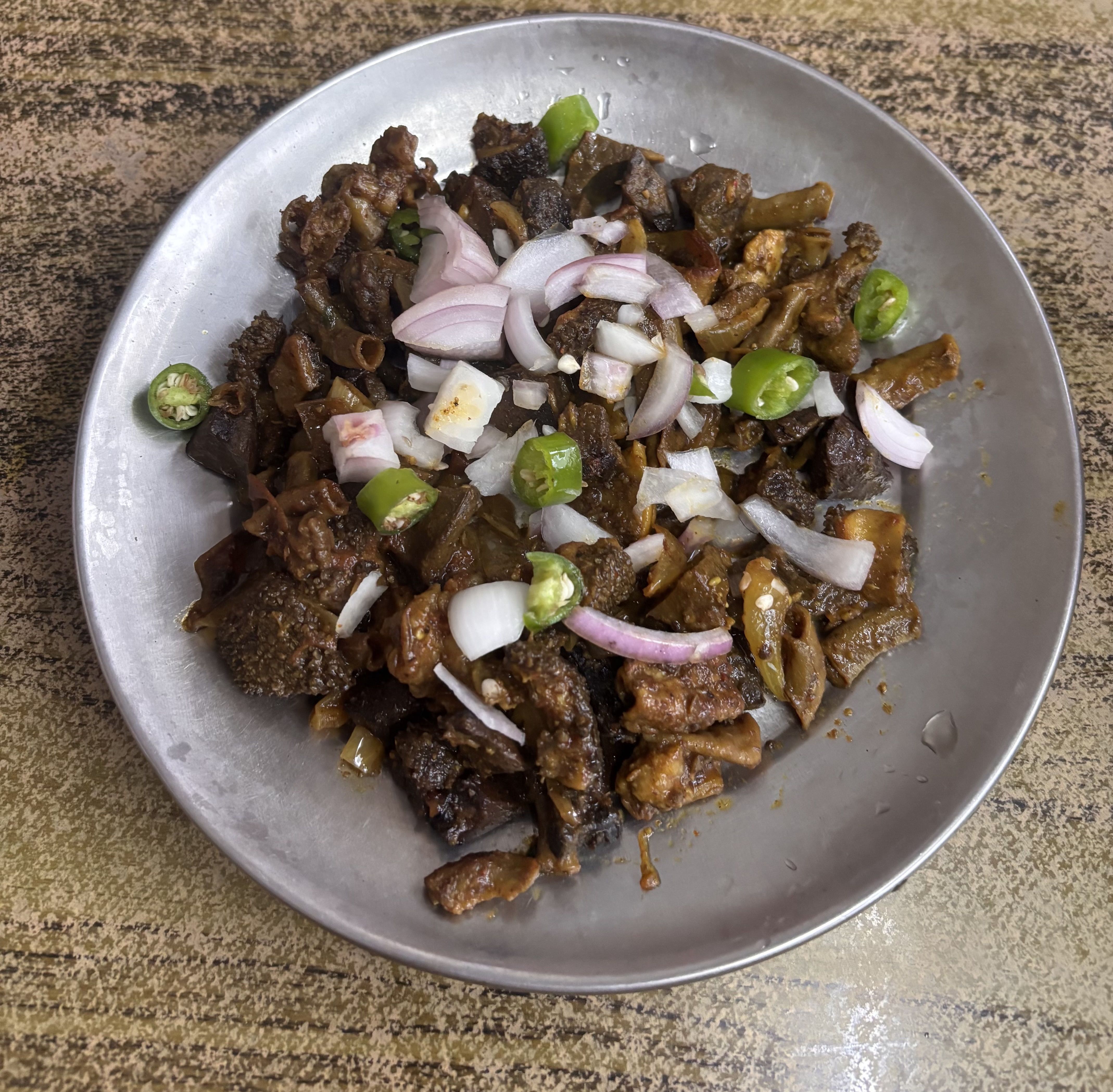 A silver plate holds a spicy dish featuring small, chopped pieces of cooked meat, likely dark and tender, mixed with various spices.
