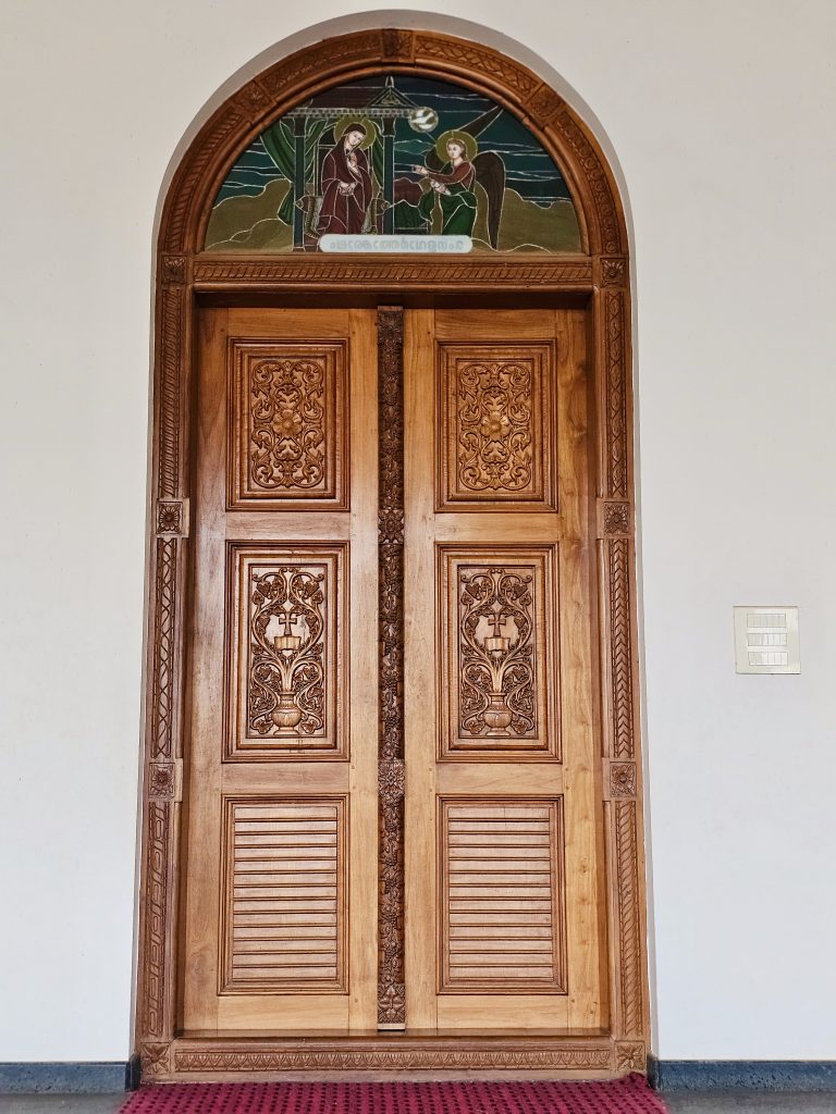 An intricately carved wooden church door with a beautiful religious artwork panel above it. The detailed woodwork and warm tones make this a strong architectural detail photograph. Location: Ramapuram, Kottayam, Kerala.