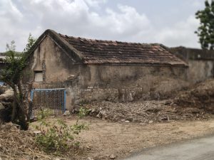 Old weathered building with a reddish-brown tiled roof, cracked walls, a blue metal gate, and a small green plant growing among the rubble.
