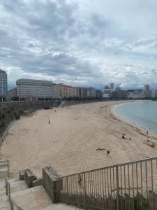 Wide view of a sandy beach with calm water, a few people, and buildings under a cloudy sky.
