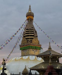 Buddhist stupa with golden eyes and colorful prayer flags against pastel clouds at sunset.
