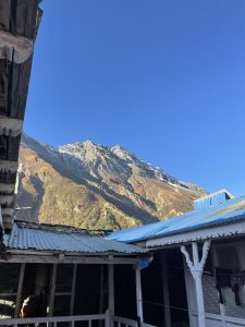 A low-angle shot looking up past the corrugated metal and blue-trimmed roofs of a wooden lodge courtyard toward a sharp, sunlit mountain peak against a clear blue sky.