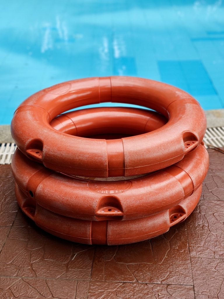 A stack of bright orange-red lifebuoys sits beside a swimming pool, with the blue water creating strong color contrast. Location: Ayamkudy, Kottayam, Kerala.