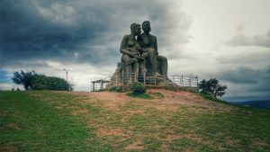 Stone sculpture of a seated family on a grassy hilltop at Ramakkalmedu, Kerala, under a cloudy monsoon sky.