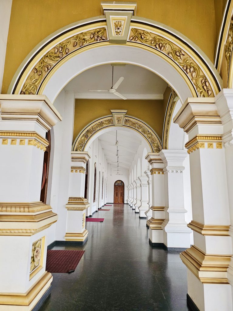 A long interior corridor with repeating white-and-gold arches, decorative columns, wooden doors, and a polished floor. The strong perspective creates an attractive sense of depth. Location: Ramapuram, Kottayam, Kerala.