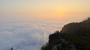 Sunrise over the clouds seen from a mountaintop. There are people at a lower level looking out at the sky.

