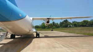 Rear view of an airplane parked on a runway. The airplane has a blue-and-silver exterior, with a prominent propeller visible on one wing. The tarmac is concrete, with a yellow line marking the runway edge. In the background, there’s a grassy area with a few scattered palm trees and what appear to be small buildings or structures. The sky is clear and bright blue, indicating a sunny day. A small vehicle, possibly a ground support cart, also appears in the distance near the airplane.
