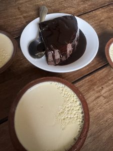 Top-down view of chocolate cake with chocolate drizzle on a white plate, served with traditional clay bowls of Juju Dhau on a wooden table.
