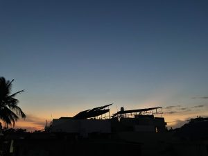 Sunset taking place while there's one star in the sky, and the colour of the sky is turning orange and yellow. Trees and terrace of a house can also be seen alongwith.