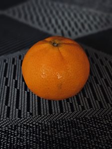A single bright orange sits against a dark textured surface. The simple composition, strong color contrast, and shallow depth of field make the fruit stand out. Location: Vazhithala, Idukki, Kerala.