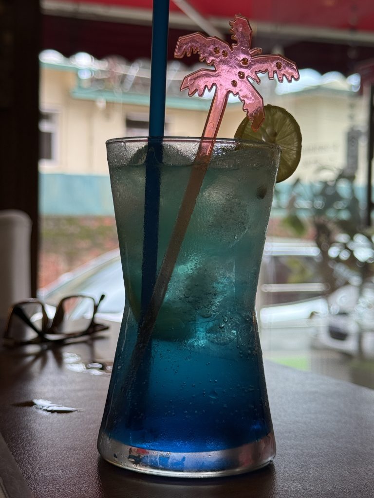 Blue iced drink with lime slice and straws in a clear glass on a wooden table.