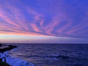 Sunset over the ocean with purple, pink, and orange skies, reflected on the water, with rocky formations along the shore.