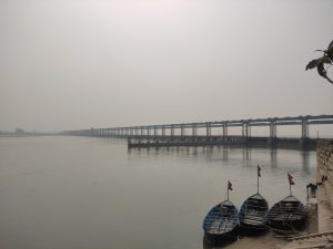 Calm Koshi River with a long bridge in the background and three small wooden boats displaying Nepali flags along the stone riverbank.