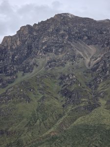 A dramatic mountain peak rises steeply with rugged, dark grey rock faces blending into sparse green alpine vegetation near its base against a cloudy sky.