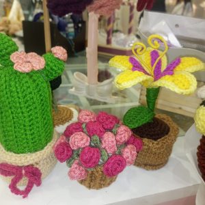 Colorful crocheted plants and flowers displayed on a table, including a small cactus with pink flowers and crocheted roses.