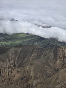A dramatic mountain ridge where heavily eroded brown rock faces give way to lush green slopes blanketed by a thick blanket of rolling white clouds.
