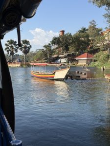 The wooden boat with a bright orange trim is moored along a calm river, suggesting it might be used for local transport or scenic cruises. Palm trees lining the riverbank indicate a warm, tropical environment. 
