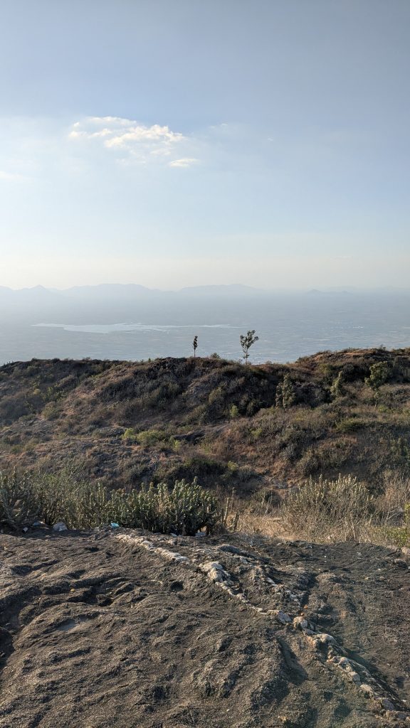 Mountain ridge with rocky terrain, grasses and shrubs in the foreground, distant hills, and a small body of water under a clear blue sky.