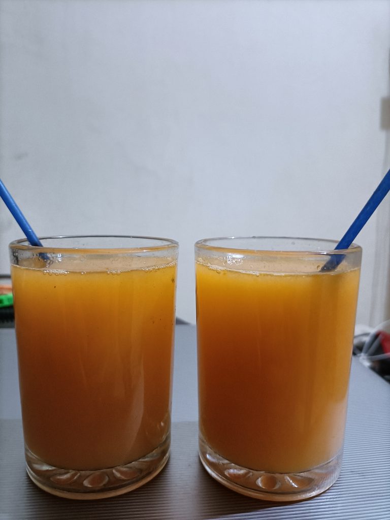 Two glasses filled with a golden-orange beverage are placed side by side on a flat surface. Both glasses are transparent with a textured base, allowing the drink's color to show through. Each glass has a blue straw. The plain, light-colored background draws attention to the drinks in the foreground.