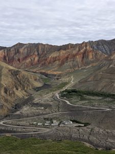 An aerial view looking down onto a winding mountain road carving through a valley floor beside a towering, multi-layered cliff with striations of red and tan rock.