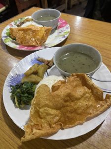 A traditional Nepali Dal Bhat meal served on white floral plates, consisting of steamed rice, crisp papadum, saag (greens), potato curry, and a bowl of lentil soup.