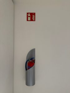Red fire extinguisher in a metal wall holder, with a red emergency sign above it.