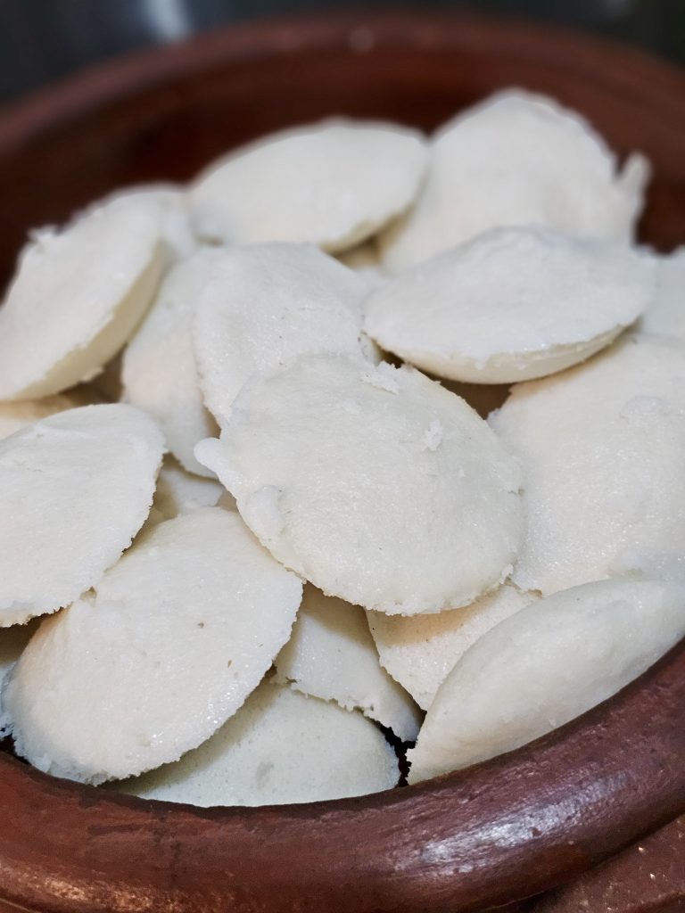 A close-up of soft idlis arranged in a traditional earthen serving bowl. The simple presentation highlights the texture and shape of this popular South Indian food. Location: Ayamkudy, Kottayam, Kerala.