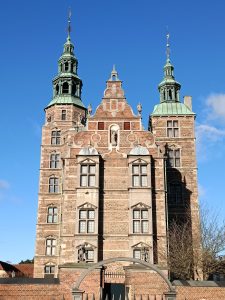 Front facade of Rosenborg Castle in Copenhagen featuring Dutch Renaissance red brick walls, sandstone decorations, and green copper spires under a clear blue sky.