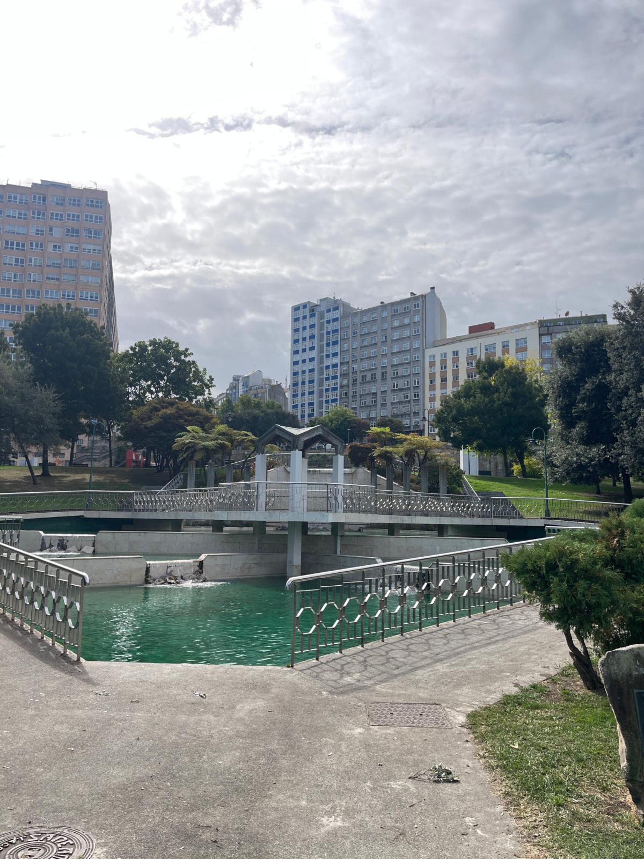An urban park with a small lake, decorative bridge, trees, and tall buildings in the background.