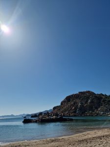 Wide-angle landscape of a sandy beach and clear sea water with a sun flare above a rocky coastal hill.