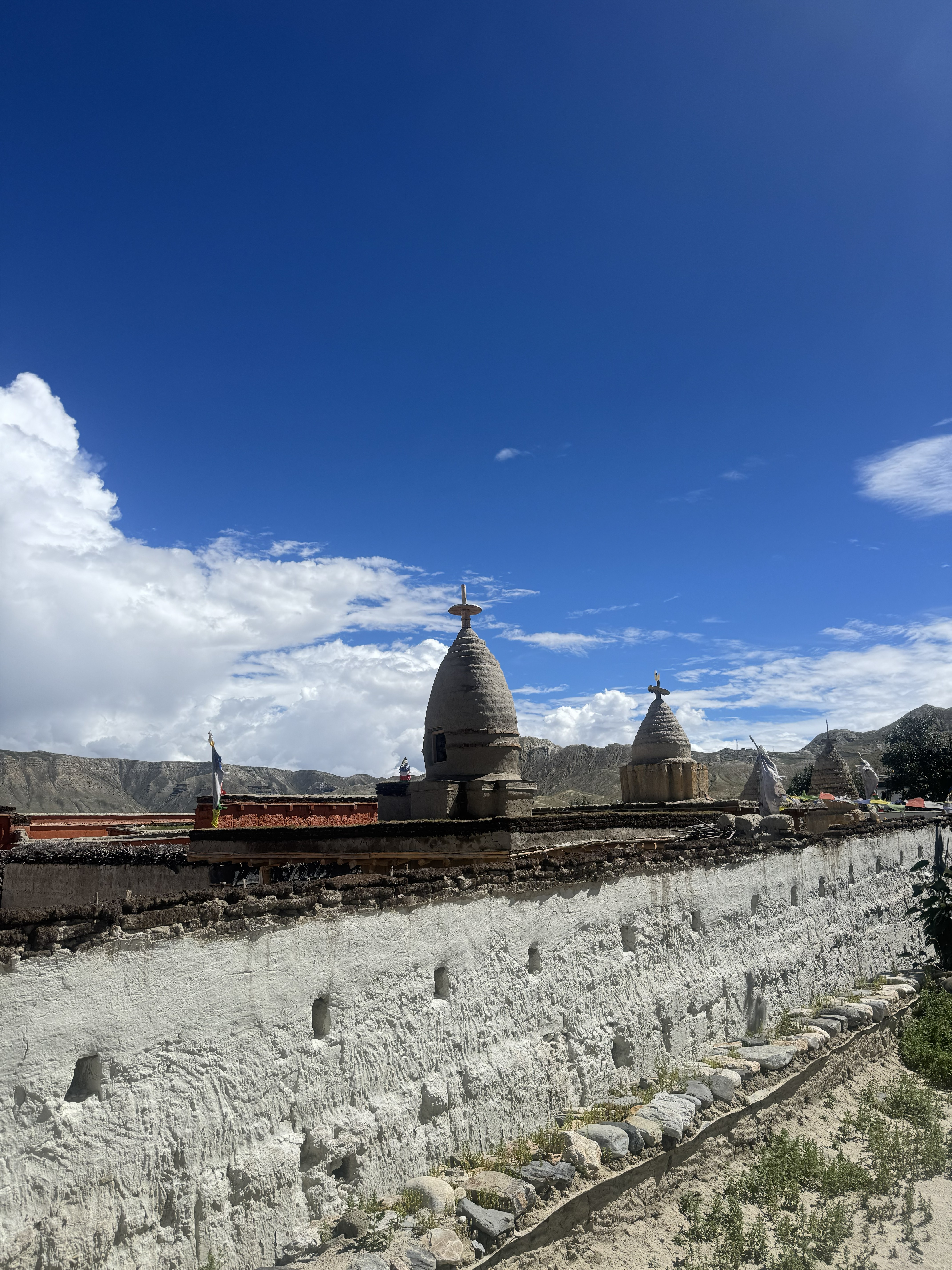 A low-angle view of traditional whitewashed earth-and-stone walls with small niches, topped by two grey, bell-shaped Buddhist stupas. Arid, mountain-ringed landscape and a vibrant blue sky with fluffy white clouds stretch behind them.