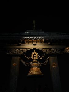 Large ornate bell hanging from a carved wooden and stone structure against a dark background.
