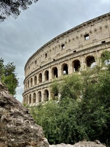 The Colosseum in Rome, Italy, is viewed from below with its arched stone facade, framed by green shrubs and a rocky outcrop in the foreground under an overcast sky.
