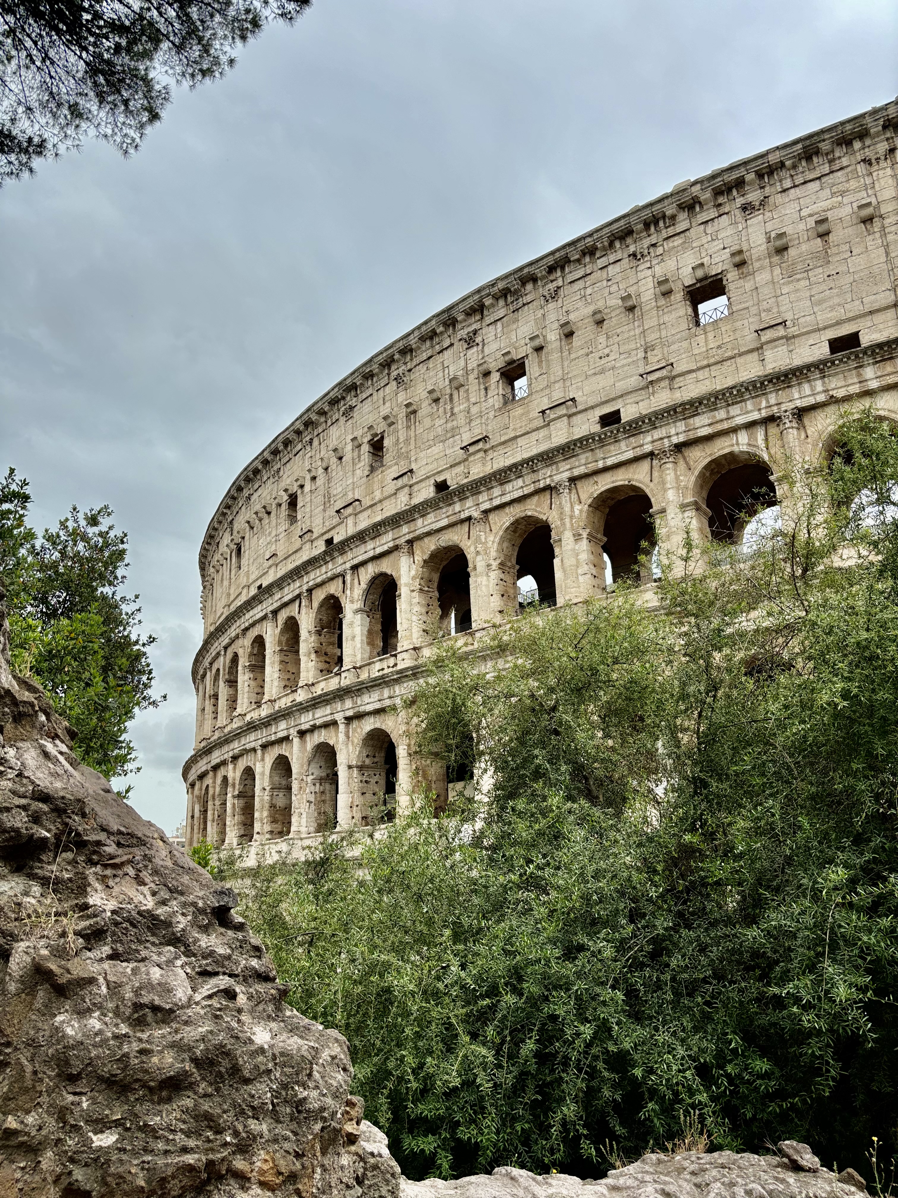 The Colosseum in Rome, Italy, is viewed from below with its arched stone facade, framed by green shrubs and a rocky outcrop in the foreground under an overcast sky.