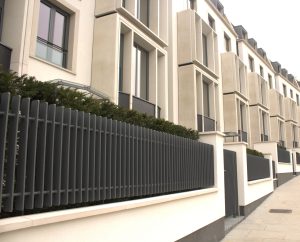 Low-angle perspective view of modern townhouse exteriors with stone window frames and dark metal slatted fencing along a sidewalk.