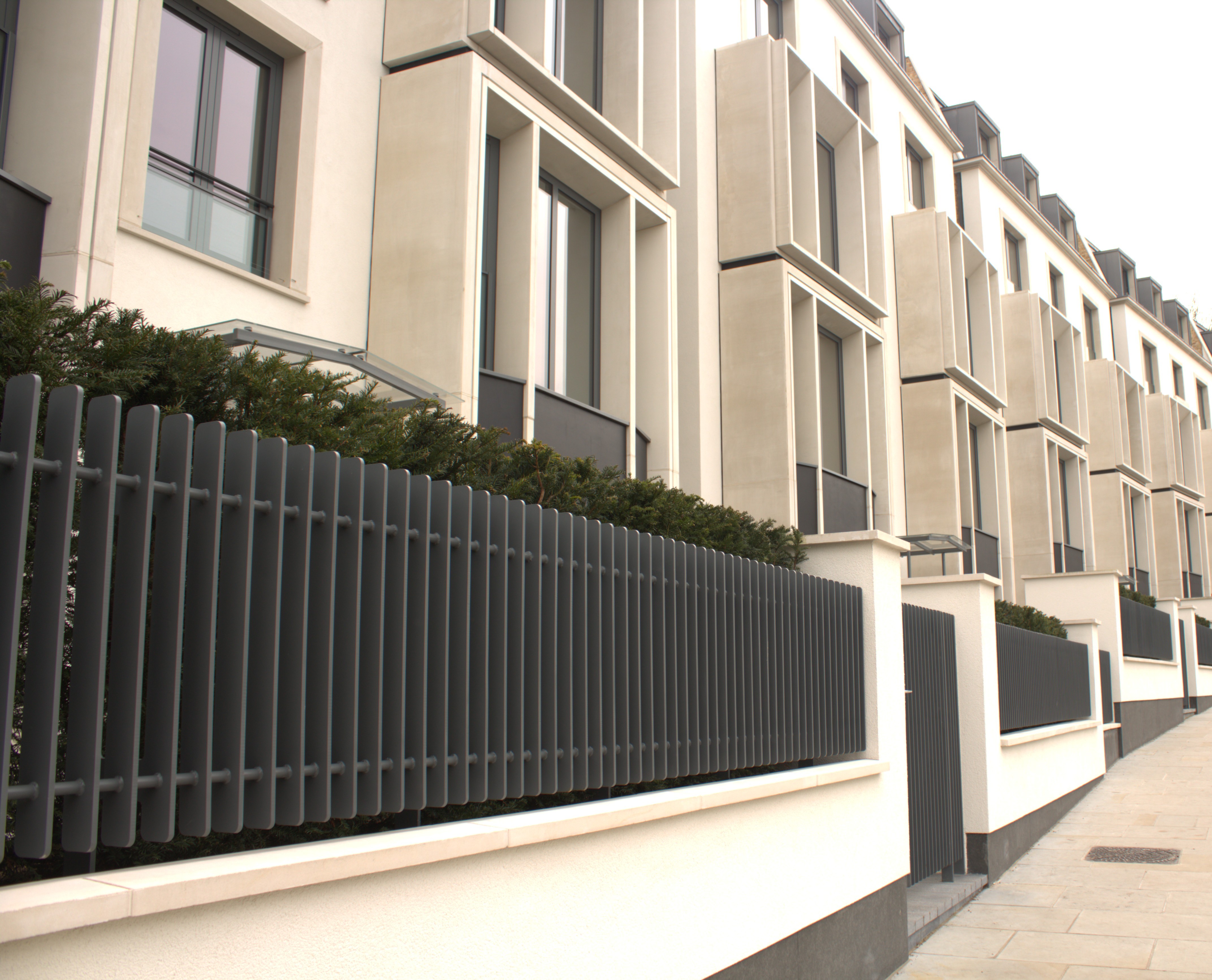 Low-angle perspective view of modern townhouse exteriors with stone window frames and dark metal slatted fencing along a sidewalk.
