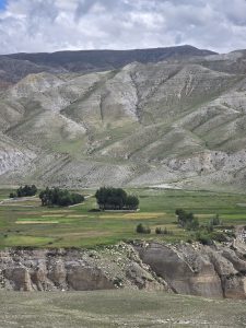 Green valley fields and trees surrounded by steep, dry rocky slopes under a cloudy sky.