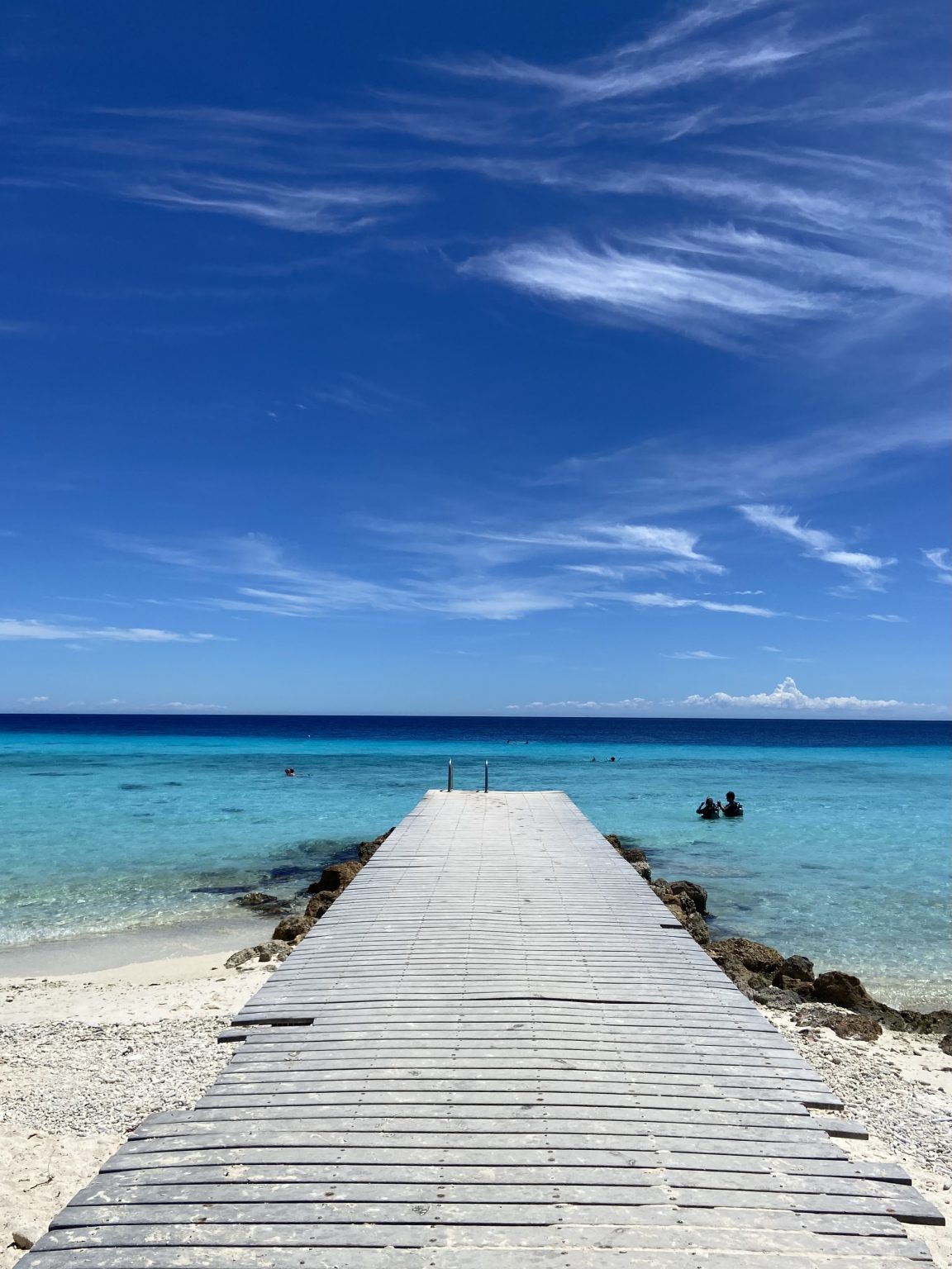 A wooden jetty running into clear turquoise sea where swimmers are out in the water under a blue sky.
