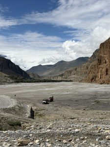 Heavy construction vehicles parked in a wide, stony riverbed surrounded by towering, dry mountain walls under a bright, partly cloudy sky.