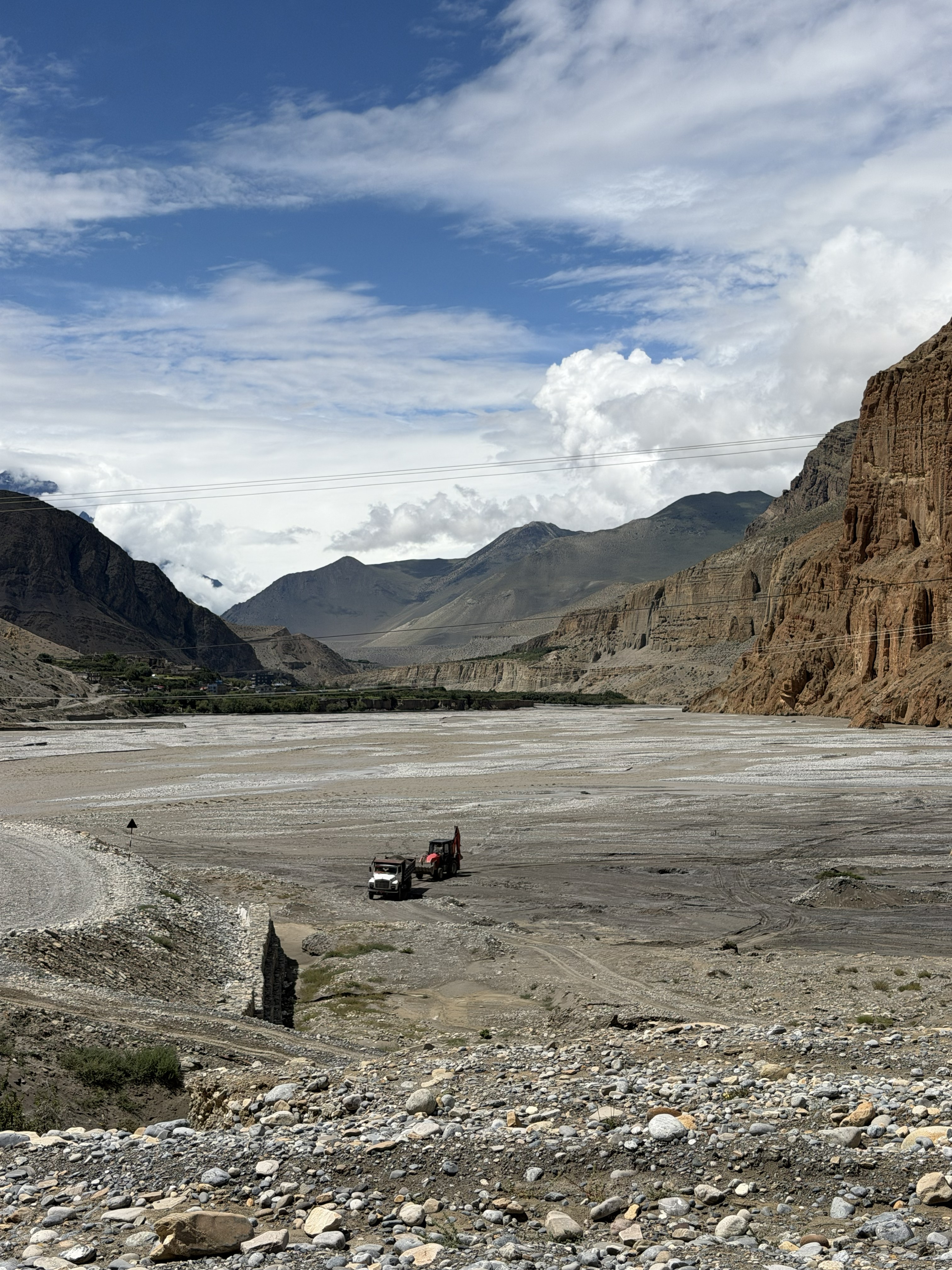 Heavy construction vehicles parked in a wide, stony riverbed surrounded by towering, dry mountain walls under a bright, partly cloudy sky.
