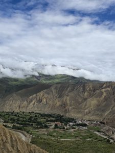 A wide elevated vista capturing a village along green valley fields, towering eroded cliffs, and rolling low clouds meeting a clear blue sky.