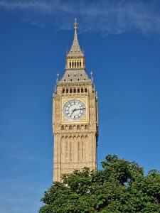Low-angle shot of the golden gothic Elizabeth Tower clock face against a clear blue sky framed by tree leaves at the bottom.