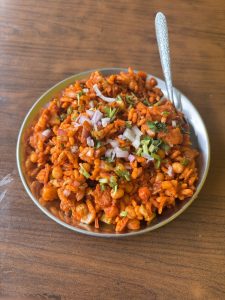 Colorful puffed rice snack with chopped onions and green herbs on a silver plate, served with a spoon on a wooden table.
