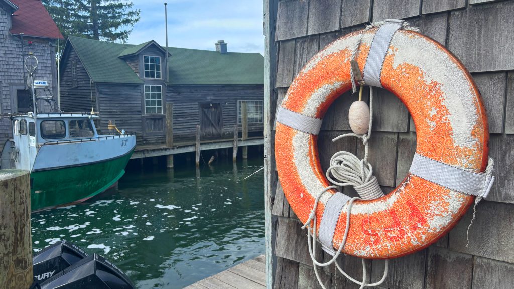 The right side of the image features a worn life ring hanging on a wall made of wooden shakes.  The left side is a narrow body of water with a fishing boat in it, and a fishing shack on the far side.